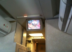 The image shows the interior of an airplane, specifically the area near the exit door. There is a small television screen mounted on the ceiling, displaying a program. The exit sign is illuminated below the TV screen. The walls and ceiling of the airplane cabin are visible, along with some overhead compartments.