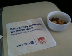 The image shows a white napkin on an airplane tray table. The napkin has text that reads, "Before they move us, we move them. Flying Team USA for over 30 years." Below the text are the logos for United Airlines and the USA Olympic Team. Next to the napkin is a small white bowl containing a mix of nuts.