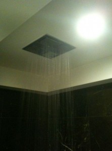 The image shows a ceiling-mounted square showerhead with water flowing from it. The showerhead is installed in a bathroom with dark tiles on the walls. A bright ceiling light is also visible near the showerhead.