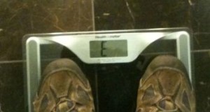 The image shows a pair of feet wearing brown shoes standing on a digital scale. The display on the scale shows the letter "E". The background appears to be a dark-colored floor.