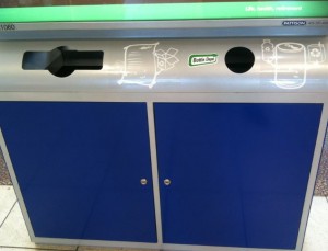 The image shows a recycling station with three separate disposal slots. Each slot is labeled with an icon indicating the type of waste it is meant for. The left slot has an icon of a hand inserting a piece of paper, the middle slot has an icon of a bottle, and the right slot has an icon of a can. The station has a blue lower section with two doors, likely for accessing the bins inside. The top section is silver with a green strip at the top.