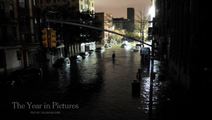 The image shows a flooded urban street at night. The street is filled with water, and a person is seen walking through the flood. Buildings line both sides of the street, and there are cars partially submerged in the water. The streetlights and some building lights are on, casting reflections on the water. The text "The Year in Pictures" and "Text by Colin McGowan" is visible in the bottom left corner of the image.