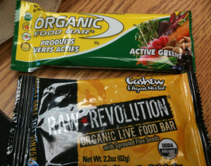 The image shows two packaged food bars. The top bar is labeled "Organic Food Bar" with "Active Greens" and "Produits Verts Actifs" written on the packaging, which is green and yellow with images of various vegetables. The bottom bar is labeled "Raw Revolution Organic Live Food Bar" with "Cashew & Agave Nectar" and "with Sprouted Flax Seeds" written on the packaging, which is yellow and black. Both bars are certified organic, as indicated by the USDA Organic seal on the bottom bar.