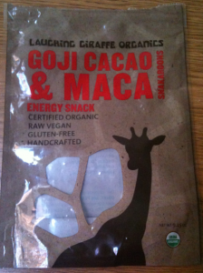 The image shows a brown packaging bag for "Laughing Giraffe Organics Goji Cacao & Maca Snakaroons." The text on the bag highlights that it is an energy snack that is certified organic, raw vegan, gluten-free, and handcrafted. The bag features a silhouette of a giraffe and has a small transparent window through which the contents can be seen. The net weight is 3.5 oz (99g), and there is an organic certification logo at the bottom right corner.