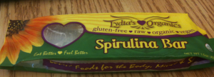 The image shows a packaged food item labeled "Spirulina Bar" from Lydia's Organics. The packaging is predominantly green and yellow with a sunflower design on the left side. The text on the package indicates that the bar is gluten-free, raw, organic, and vegan. The slogan "Eat Better, Feel Better" is also visible on the packaging. The package is placed on a wooden surface.