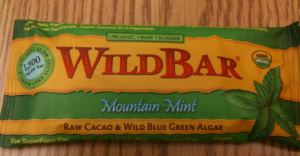 The image shows a green and yellow wrapper of a product called "WildBar." The flavor indicated on the wrapper is "Mountain Mint." The wrapper also mentions that the bar contains "Raw Cacao & Wild Blue Green Algae" and is labeled as "Organic," "Raw," "Kosher," "Gluten Free," and "Vegan." There is an image of mint leaves on the right side of the wrapper.