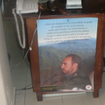 The image shows a wooden desk with a telephone and some other items on top. Below the desk, there is a poster featuring a man in a military uniform with a beard, speaking into a microphone. The background of the poster shows a mountainous landscape. The poster also contains text in Spanish.