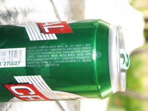 The image shows a green aluminum can of beer lying on its side. The can has a red and white label with the word "CR" visible, likely part of the brand name. There is a barcode and some text in Spanish on the can, which includes instructions and ingredients. The can is placed on a surface with a blurred background, possibly outdoors.