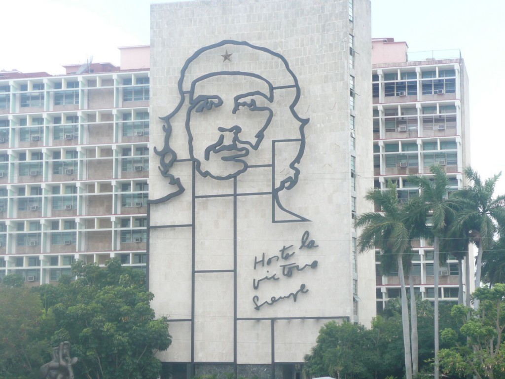 The image shows a large building with a prominent mural of Che Guevara's face on its facade. The mural is a black outline of his face, including his iconic beret with a star. Below the mural, there is a cursive inscription that reads "Hasta la victoria siempre." The building has multiple windows and is surrounded by trees and palm trees.