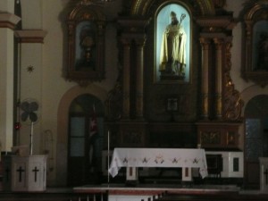 The image shows the interior of a church. At the center is an altar covered with a white cloth adorned with crosses. Behind the altar, there is a large, ornate structure with a statue of a religious figure illuminated in a niche. The surrounding architecture includes arches and decorative elements. The overall lighting is dim, with the statue being the focal point due to its illumination.