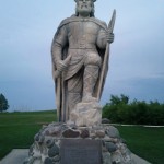 A large stone statue of a Viking warrior stands on a pedestal made of rocks. The Viking is depicted wearing a helmet with horns, a cloak, and armor, and is holding a sword in one hand and a shield in the other. The statue is set outdoors on a grassy area with some trees and bushes in the background. The sky is overcast. There is a plaque on the pedestal, but the text is not legible in the image.