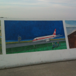 The image shows a mural painted on a concrete wall. The mural consists of three distinct sections. The middle section features an airplane with "Air Canada" written on it, taxiing on a runway with a control tower in the background. There are also a few people depicted near the fence in the foreground. The left section of the mural shows a whimsical scene with two cartoonish characters standing under a tree, and the right section appears to be an abstract or partially completed painting with a brown and blue color scheme.