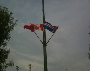 The image shows two flags on a flagpole against a cloudy sky. The flag on the left is the national flag of Canada, featuring a red maple leaf in the center with red borders on either side. The flag on the right is the national flag of Iceland, which has a blue background with a white cross and a red cross inside the white cross. There are trees partially visible on the left and right sides of the image.