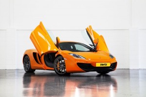 Alt text: An orange sports car with its butterfly doors open, parked indoors on a shiny floor. The car has a sleek design and a Hertz logo on the front license plate. The background is a plain white wall.