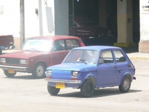 The image shows two small cars parked on a street. The car in the foreground is blue, while the car in the background is red. Both cars have yellow license plates. The background includes a building with an open garage or entrance.