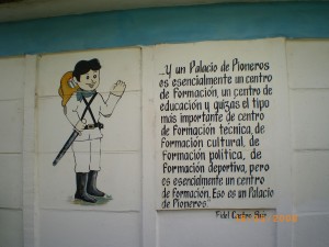 The image shows a mural on a wall. On the left side, there is a cartoon drawing of a child dressed in a white uniform with a red scarf, black boots, and a black hat, holding a flag. The child is smiling and waving. On the right side, there is a block of text in Spanish that reads:

"Y un Palacio de Pioneros es esencialmente un centro de Formación, un centro de educación y quizás el tipo más importante de centro de Formación técnica, de Formación cultural, de Formación política, de Formación deportiva, pero es esencialmente un centro de Formación. Eso es un Palacio de Pioneros."

The text is attributed to "Fidel Castro Ruz 2003" at the bottom right corner.