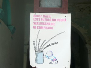 The image shows a poster hanging on a wall. The poster has text in Spanish that reads: "Señor Bush: Este pueblo no podrá ser engañado, ni comprado." Below the text, there is an illustration of a child throwing a piece of paper into a trash can. The trash can has words coming out of it, including "Transición" and "Becas para jóvenes." The background of the image appears to be an old, possibly abandoned building with peeling paint and an arched doorway.