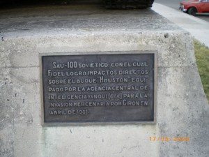 A plaque mounted on a concrete structure with Spanish text that reads: "SAU-100 soviético, con el cual Fidel logró impactos directos sobre el buque 'Houston' equipado por la Agencia Central de Inteligencia Yanqui (CIA) para la invasión mercenaria por Girón en abril de 1961." The date "17/04/2008" is visible in the bottom right corner of the image. In the background, there is a red car parked on a road.