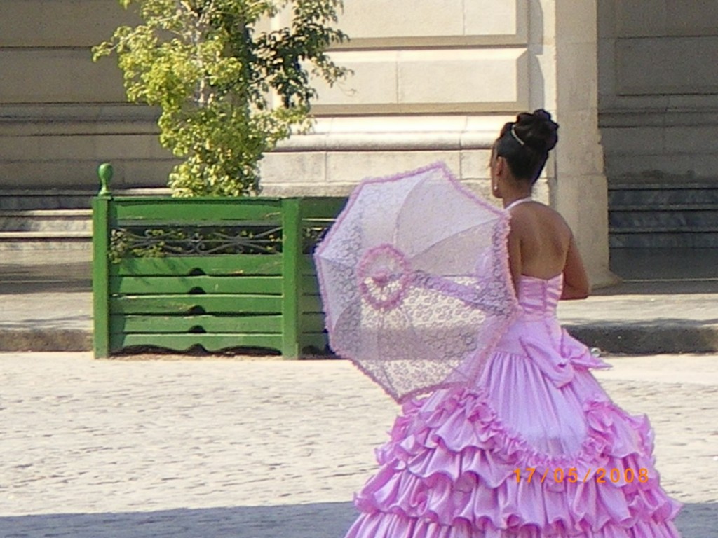 A person is seen from behind, wearing a pink ruffled dress and holding a pink lace parasol. The person has their hair styled in an updo. They are standing on a cobblestone surface with a green wooden structure and some greenery in the background. The date "17/05/2008" is visible in the bottom right corner of the image.