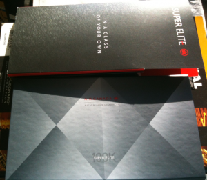 The image shows two booklets or folders. The top one is black with the text "IN A CLASS OF YOUR OWN" and "SUPER ELITE" along with a logo on the right side. The bottom one is gray with a geometric pattern and the text "AIR CANADA" and "SUPER ELITE" in the center. Both items appear to be related to a premium or elite membership program.