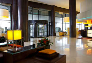 The image shows a modern hotel lobby with a sleek and contemporary design. The space features large floor-to-ceiling windows, allowing natural light to flood in. There are several tall, dark columns and a revolving glass door at the entrance. The lobby is furnished with comfortable seating areas, including cushioned benches with orange and brown pillows. There are also decorative plants and modern table lamps with yellow shades, adding a warm ambiance to the space. The floor is made of polished, light-colored tiles, and there are various lighting fixtures hanging from the ceiling.